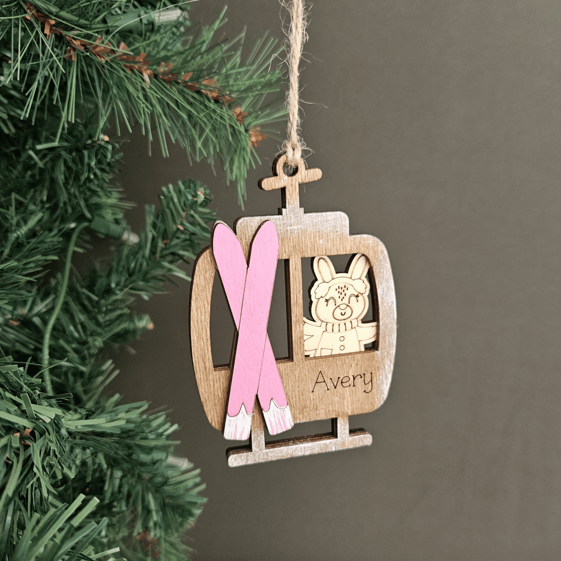 Wooden ornament shaped like a ski lift with a pink ribbon and 'Avery' engraved, hanging on a Christmas tree.