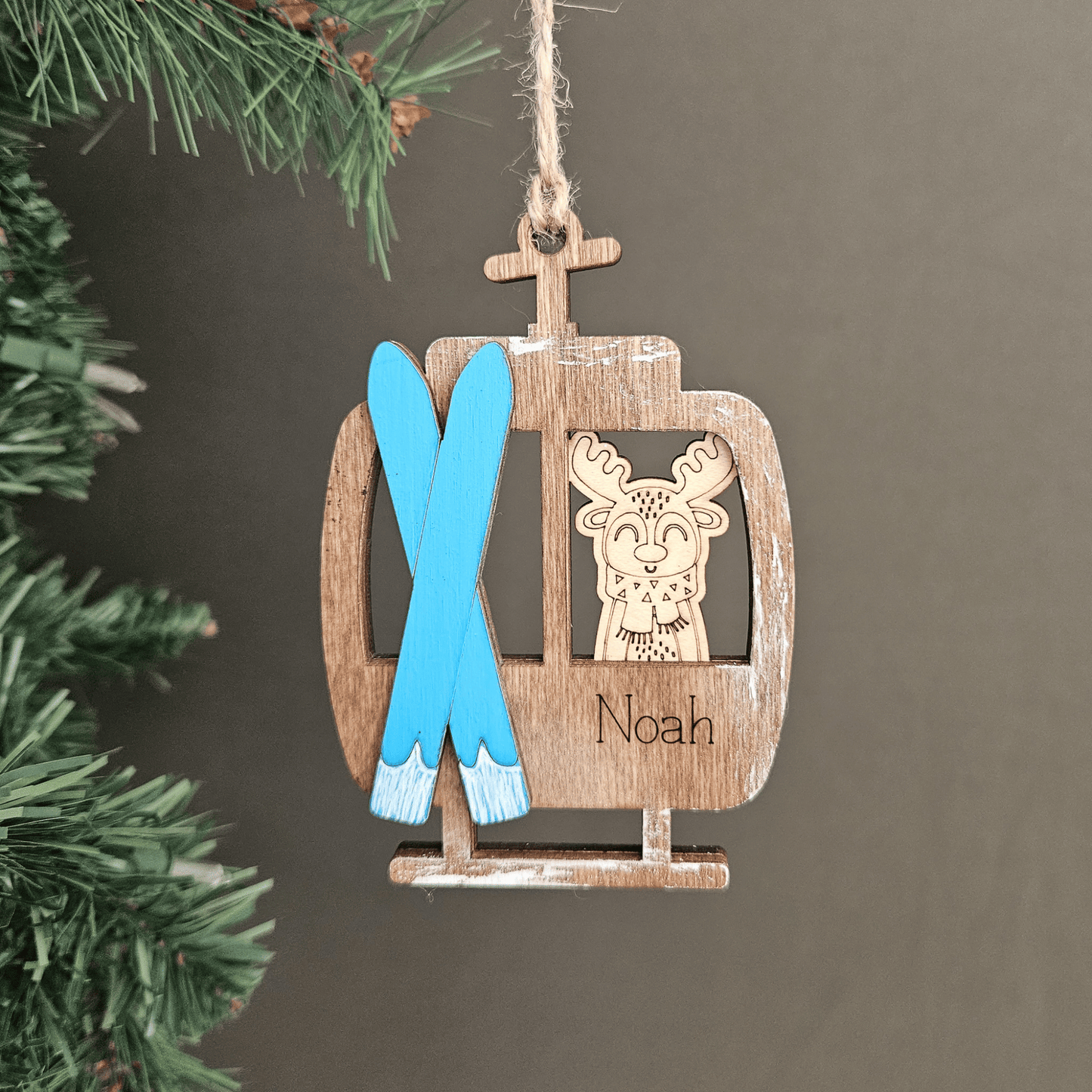 Wooden ornament shaped like a ski lift with blue skis and a reindeer design, hanging on a tree.